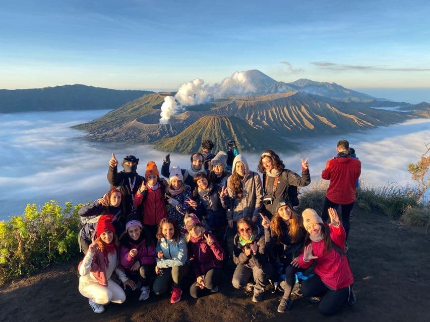 Eine WeRoad-Gruppe posiert für ein Foto auf einem Aussichtspunkt mit Blick auf einen rauchenden Vulkankrater, der über den Wolken thront.