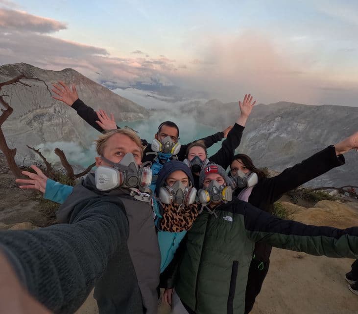Eine WeRoad-Gruppe macht ein Selfie mit Gasmasken am Rand eines Vulkankraters, darunter ein See.