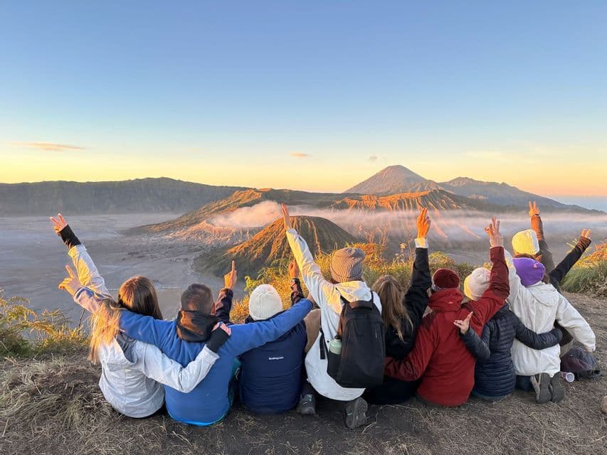 Eine WeRoad-Gruppenreise sitzt auf einem Aussichtspunkt mit erhobenen Armen und beobachtet, wie der Sonnenaufgang einen Vulkankrater beleuchtet.