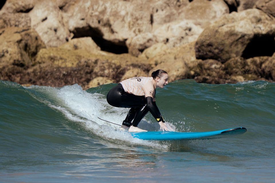 Una donna in muta da surf si accovaccia su una tavola blu, cavalcando un'onda nell'oceano con la costa rocciosa sullo sfondo.