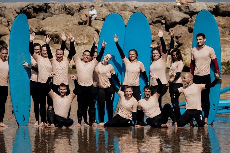 Un viaggio di gruppo WeRoad in mute da surf in posa con tavole da surf blu nell'acqua bassa di una spiaggia rocciosa.