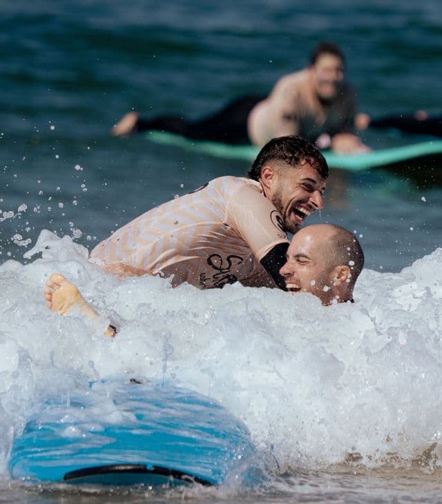 Due uomini di un viaggio di gruppo WeRoad ridono mentre cadono tra le onde dell'oceano accanto a una tavola da surf blu, con un'altra persona sullo sfondo.
