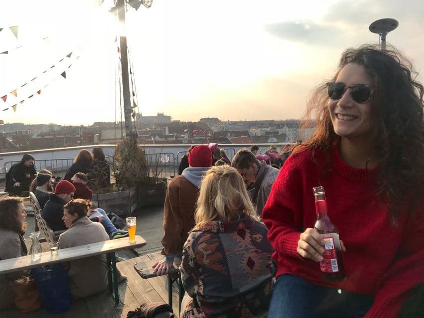 Una donna con un maglione rosso, partecipante a un viaggio di gruppo WeRoad, sorride mentre sorseggia un drink su un affollato rooftop bar al tramonto.