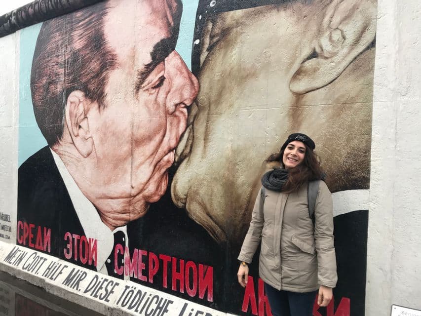Una donna con cappotto invernale e berretto sorride davanti a un grande murale su un muro di cemento raffigurante due uomini che si baciano.