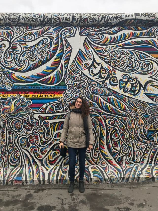 Una donna con un cappotto invernale sorride mentre posa per una foto davanti a un grande murale colorato su un muro esterno.