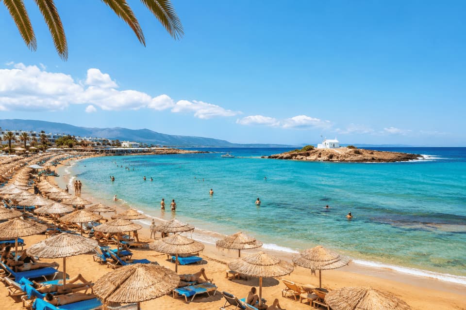 Una vista dall'alto di una spiaggia sabbiosa affollata con ombrelloni di paglia, persone che nuotano in acque turchesi e una piccola isola al largo.