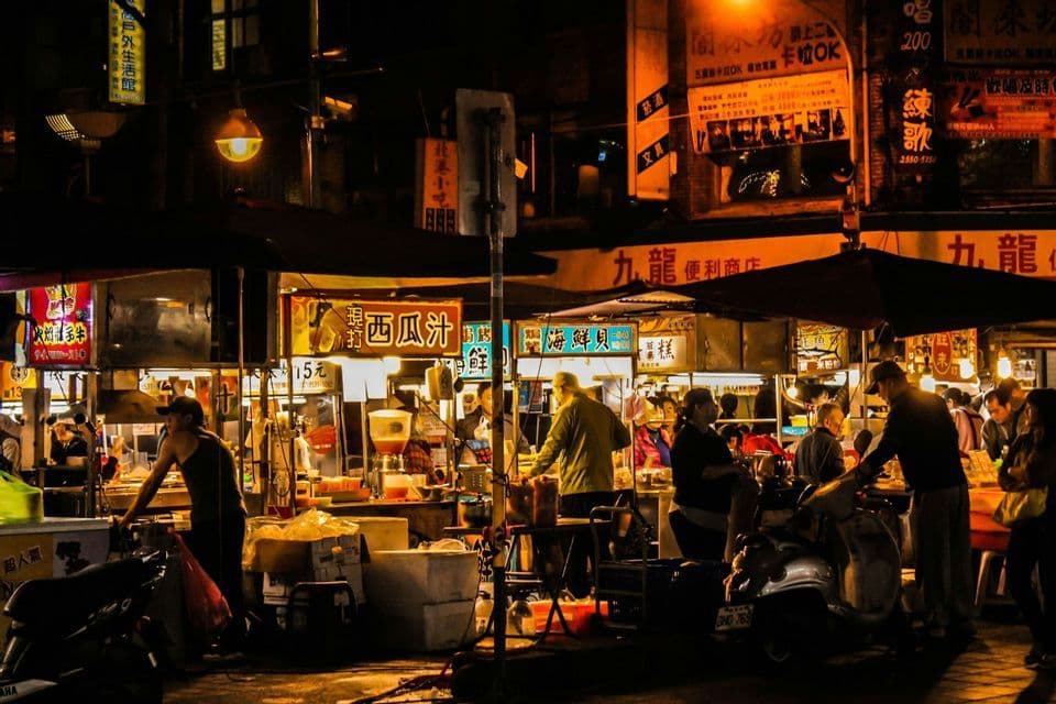 Un vivace mercato notturno asiatico con persone riunite presso bancarelle di cibo illuminate sotto insegne luminose.