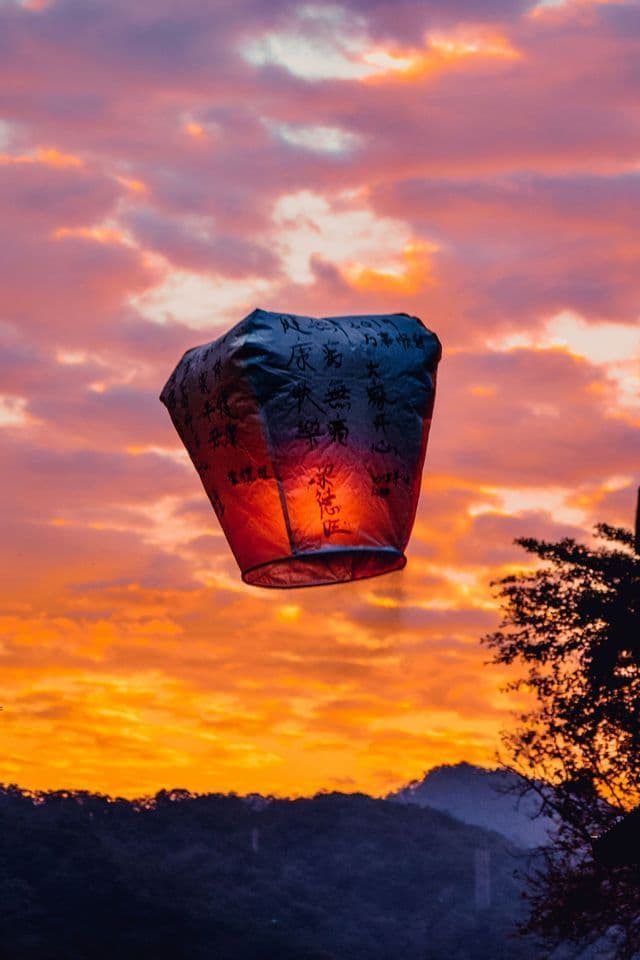 Una lanterna di carta accesa con calligrafia cinese galleggia in un cielo nuvoloso e colorato al tramonto sopra colline in silhouette.