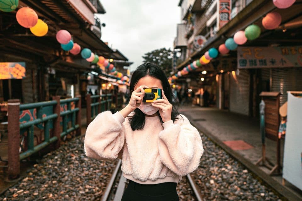 Una donna con una maschera scatta una foto sui binari di una ferrovia che attraversano un mercato di strada decorato con lanterne di carta colorate.
