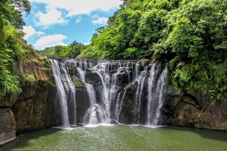 Una cascata ampia e a gradoni si getta da scure sporgenze rocciose in una pozza verde, circondata da una foresta lussureggiante sotto un cielo azzurro.