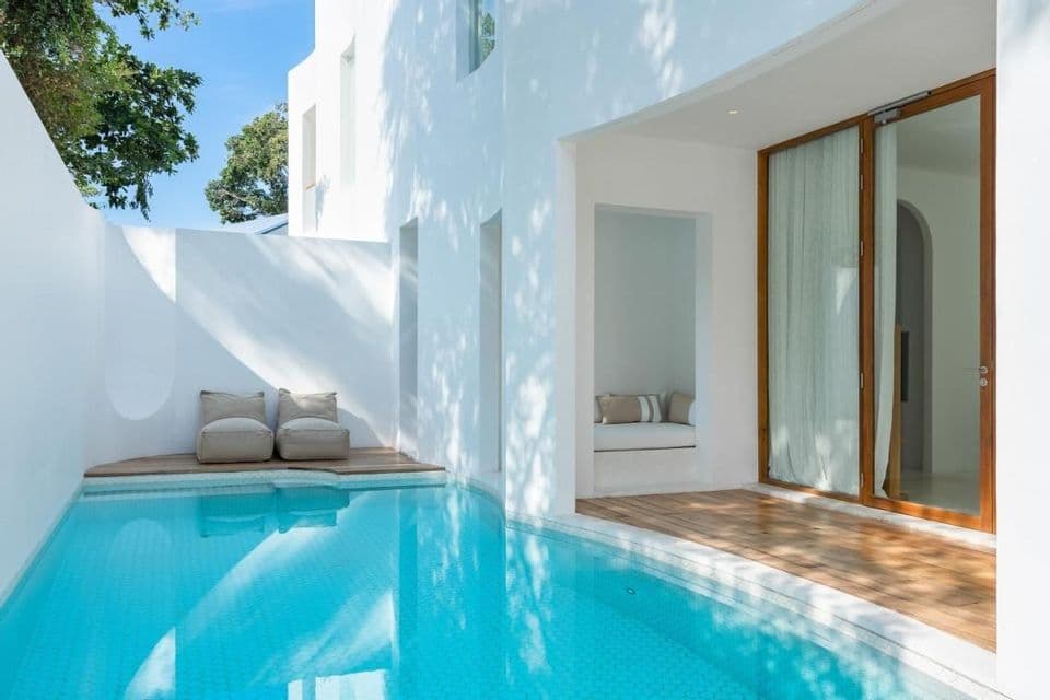 Una piscina privata soleggiata e un ponte in legno adiacenti a un moderno edificio bianco con sedute minimaliste.