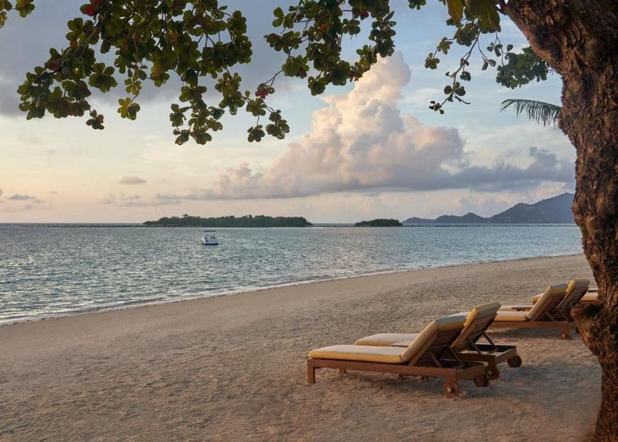 Des transats vides reposent sur une plage de sable face à la mer, au crépuscule, avec un bateau et des îles visibles au loin sous un ciel nuageux.