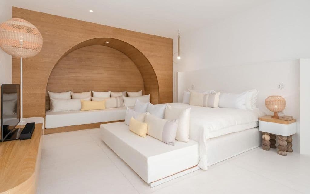 Una camera da letto bianca minimalista con un ampio letto e una parete ad arco in legno con un divano integrato, decorata con cuscini chiari.