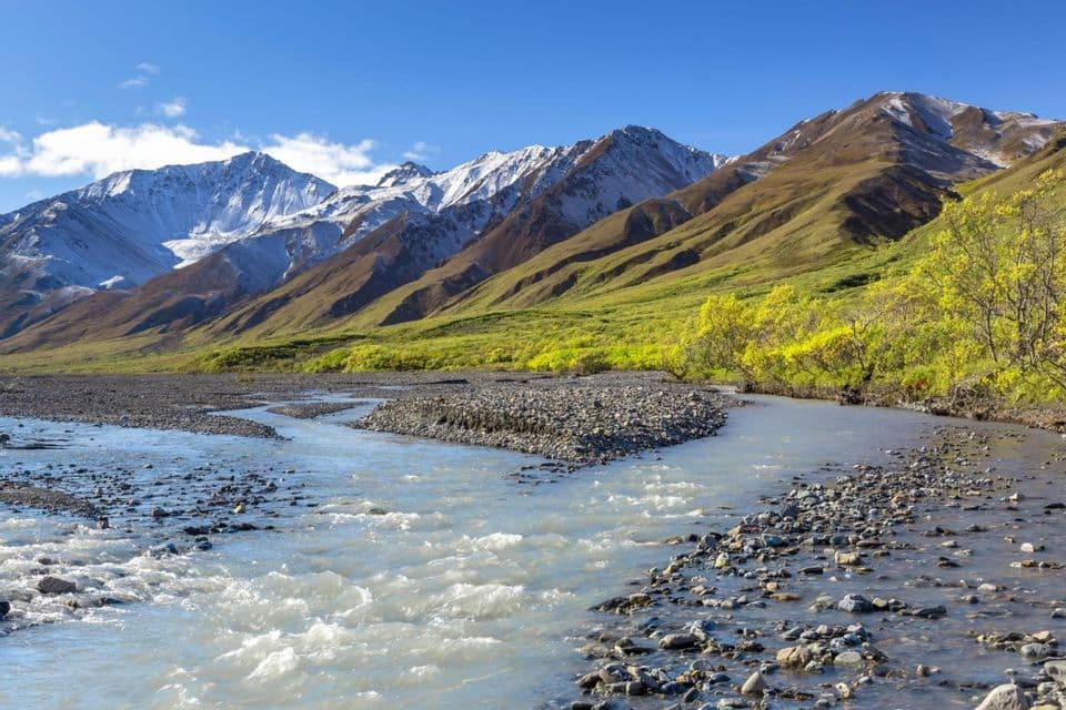 Un fiume roccioso scorre in una valle con verdi colline che conducono a montagne innevate sotto un cielo azzurro.