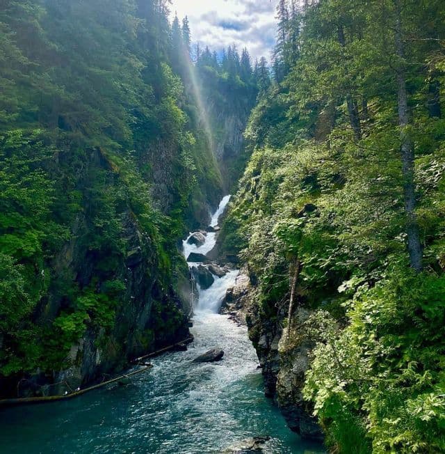 Una cascata precipita lungo una gola stretta e rocciosa, fiancheggiata da fitti alberi verdi, sfociando in un fiume turchese sottostante.
