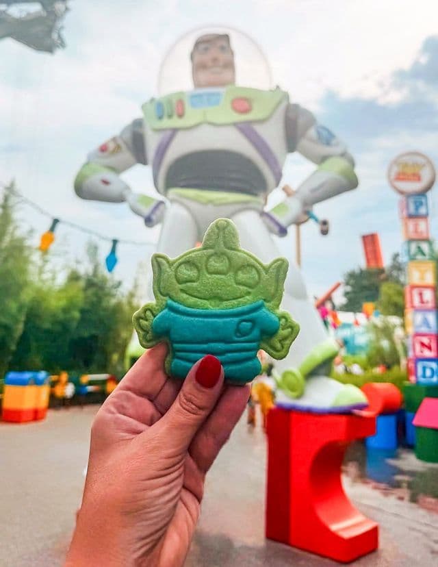 Una mano con smalto rosso tiene un biscotto verde a forma di alieno davanti a una grande statua sfocata di Buzz Lightyear.