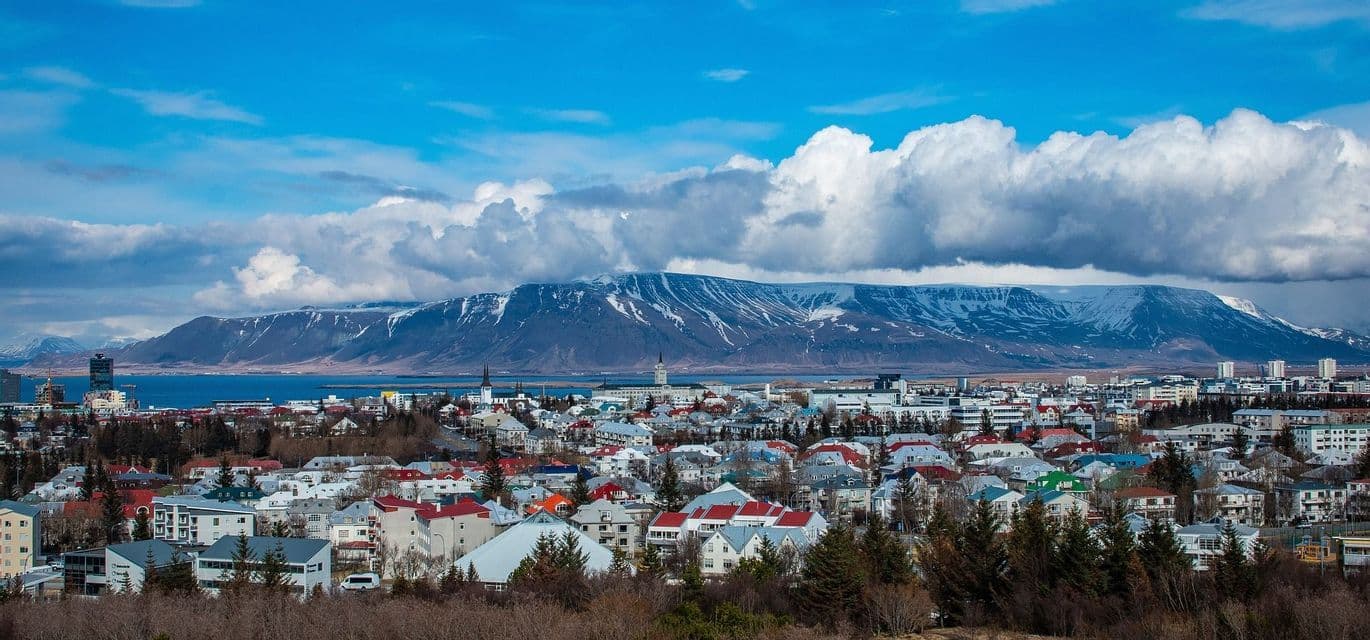 A city with colorful buildings stretches along the coast at the foot of a large, snow-capped mountain under a partly cloudy sky.