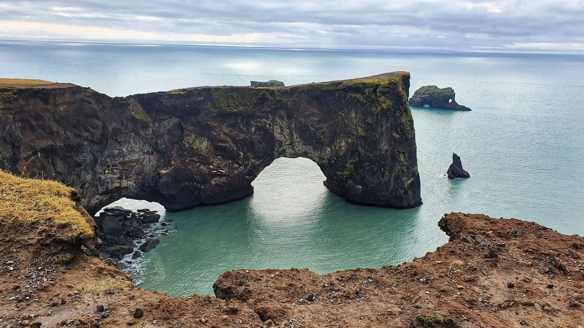 View from a grassy cliff of a large natural rock arch extending into a turquoise ocean with sea stacks in the background.