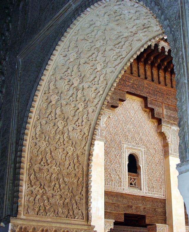 Un arco ornamentado con detalladas tallas en piedra enmarca la vista de una pared interior decorada y una pequeña ventana.