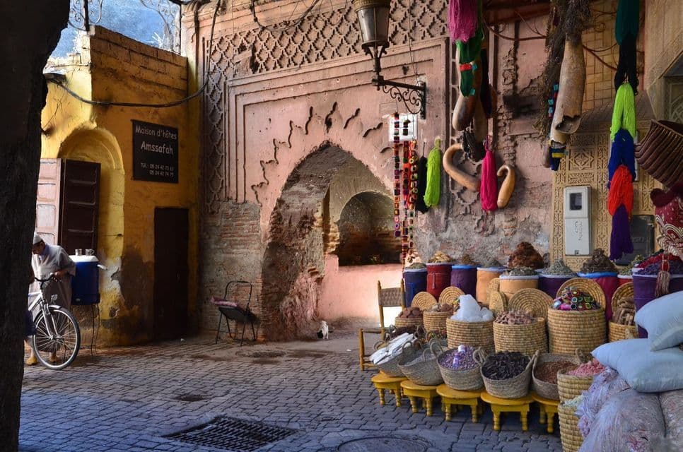 Un puesto de especias con especias de colores en cestas tejidas, situado en un estrecho callejón empedrado junto a un arco decorativo.
