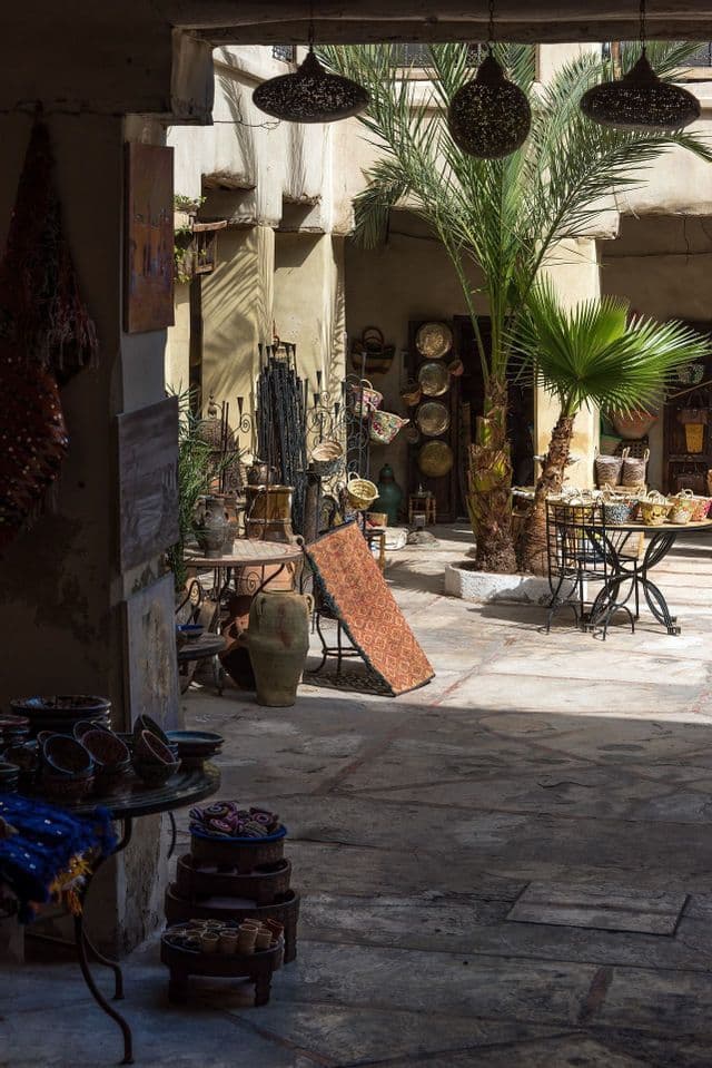 Un patio soleado de un mercado con una palmera, exhibiendo artesanías como cerámica, alfombras y faroles colgantes.