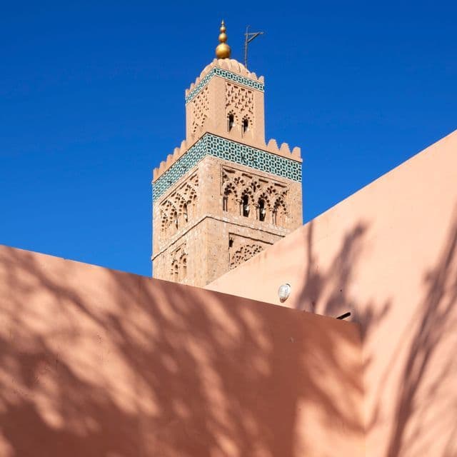 Un minarete de piedra ornamentado con azulejos verdes se eleva contra un cielo azul profundo, visto sobre un muro color salmón con sombras de palmeras.