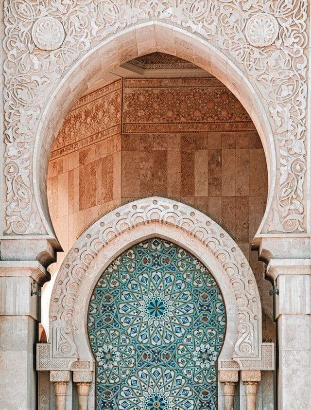 Primer plano de un arco morisco de piedra intrincadamente tallado que enmarca un arco más pequeño con mosaicos geométricos azules y verdes.