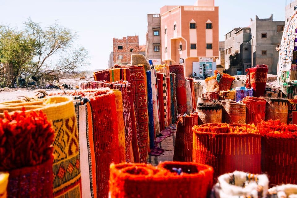 Alfombras enrolladas coloridas y cestas tejidas se exhiben para la venta en un mercado al aire libre con edificios al fondo.
