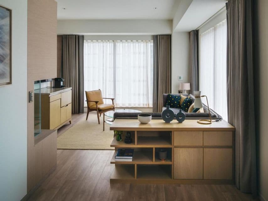 Un salon moderne avec des meubles en bois clair, un canapé gris et de grandes fenêtres avec des voilages et des rideaux épais.