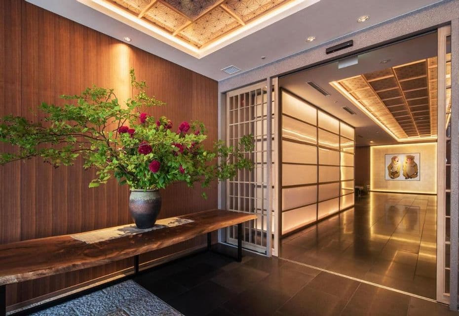 Ein großes Blumenarrangement in einer Keramikvase steht auf einer Holzbank in einer Lobby im japanischen Stil neben einem warm beleuchteten Flur.