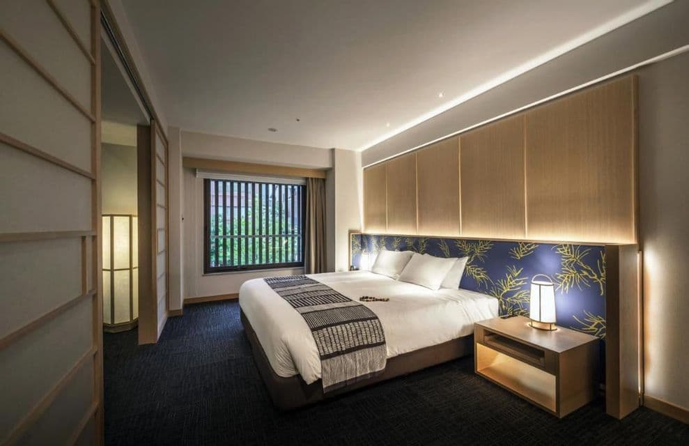 Intérieur d'une chambre d'hôtel moderne avec des éléments de design japonais, incluant un grand lit, un paravent shoji et une fenêtre avec des stores en bois.