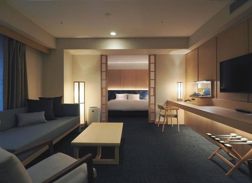 Una suite de hotel cálidamente iluminada con decoración moderna de estilo japonés, que cuenta con una sala de estar y un dormitorio separado visible a través de una amplia entrada.