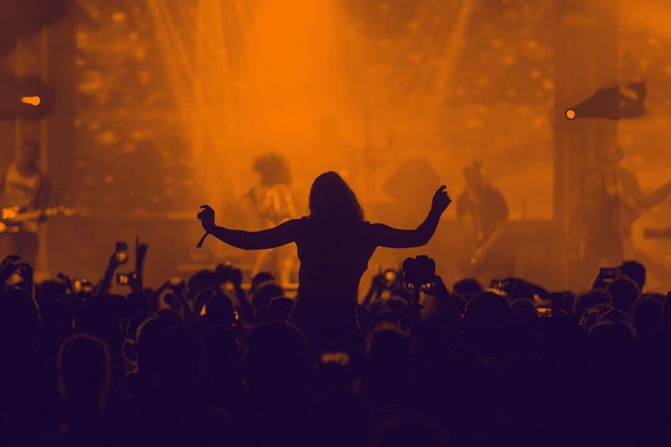 Une personne assise sur les épaules de quelqu'un, les bras levés, dans une foule lors d'un concert sous des lumières de scène oranges.