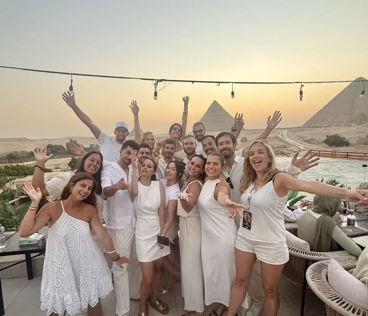 Un groupe WeRoad vêtu de blanc pose pour une photo sur une terrasse avec les pyramides de Gizeh en arrière-plan au coucher du soleil.