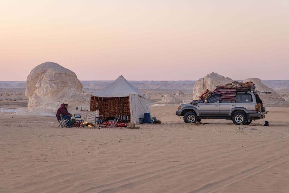 Un voyage de groupe WeRoad en camping près d'un feu dans le désert, à côté de leur tente et d'un SUV, avec de grandes formations rocheuses blanches en toile de fond.