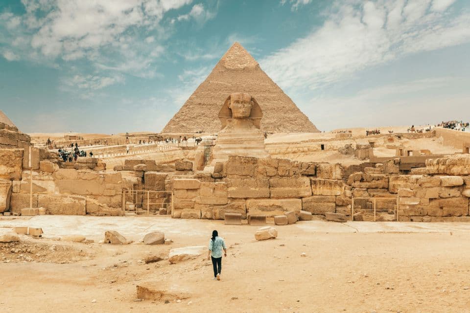 Une personne de dos marche sur le sable en direction du Grand Sphinx, avec une pyramide en arrière-plan sous un ciel nuageux.