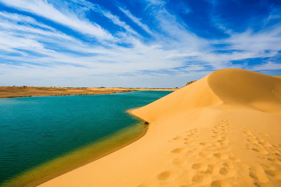 Une dune de sable doré avec des empreintes de pas descend vers une étendue d'eau turquoise dans un désert sous un ciel bleu nuageux.