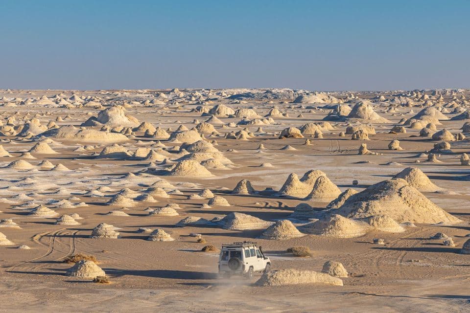 Un véhicule tout-terrain blanc roule dans un paysage désertique parsemé de formations rocheuses blanches et calcaires sous un ciel dégagé.