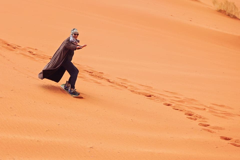 Une personne en robe et lunettes de soleil descend en sandboard une vaste dune de sable orange dans le désert.