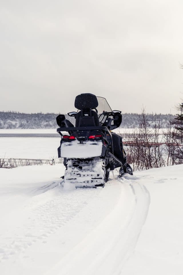 Una motoslitta nera è parcheggiata nella neve profonda, lasciando tracce dietro di sé, con un lago ghiacciato e alberi sullo sfondo.