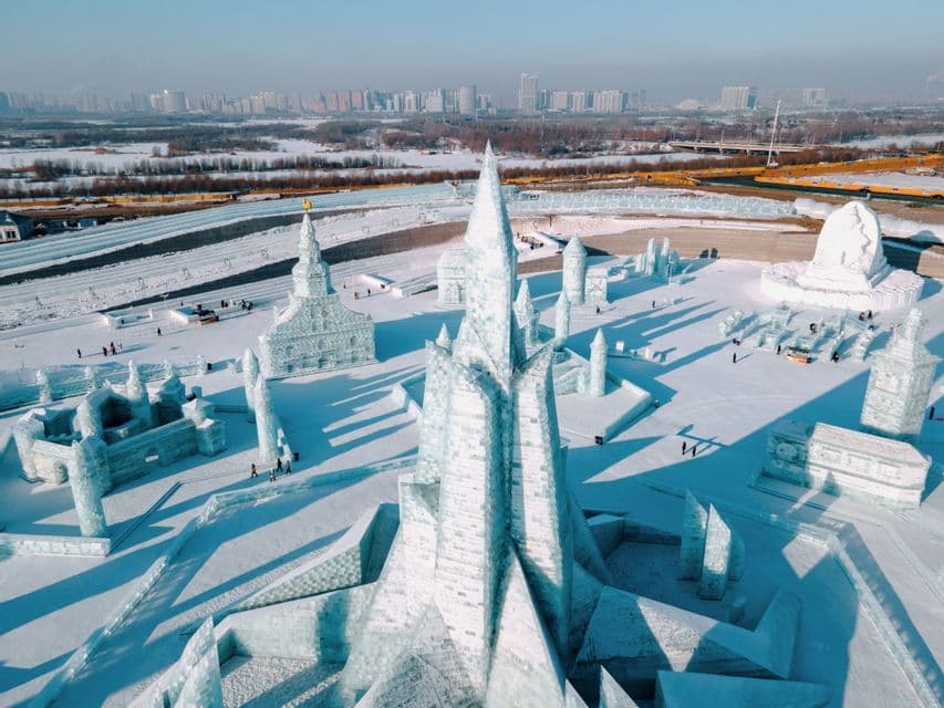 Una vista aerea di enormi sculture di ghiaccio e castelli in un paesaggio innevato, con uno skyline di città distante sotto un cielo limpido.