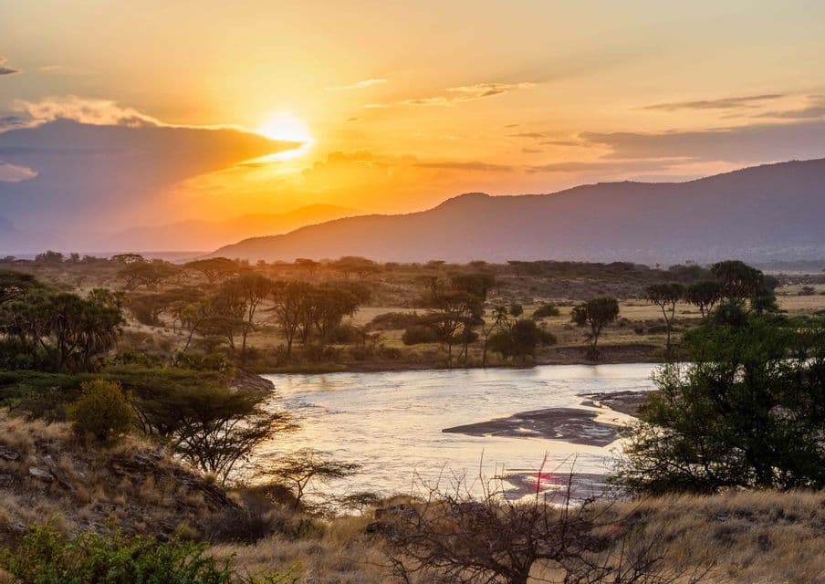 The sun sets over a wide river flowing through a savanna landscape with acacia trees and distant mountains.