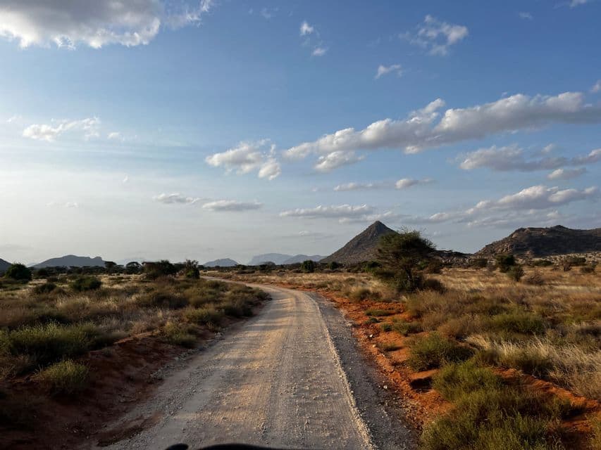 Eine gewundene Schotterstraße zieht sich durch eine trockene Savannenlandschaft mit einem kegelförmigen Berg unter einem teilweise bewölkten blauen Himmel.