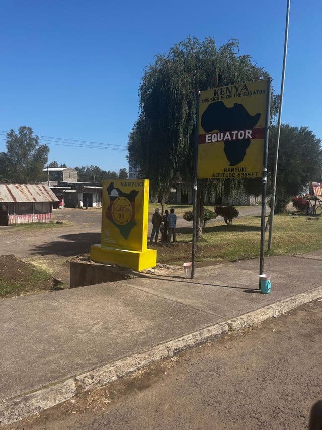 Two large yellow signs marking the equator line in Nanyuki, Kenya, stand by a road with a few people nearby under a clear sky.