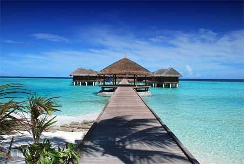 A wooden pier extends over turquoise water towards thatched-roof bungalows under a deep blue sky.