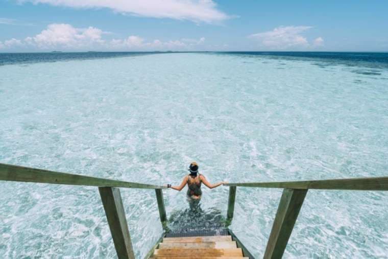 A person viewed from behind descends wooden stairs into clear, shallow turquoise water under a blue sky with clouds.