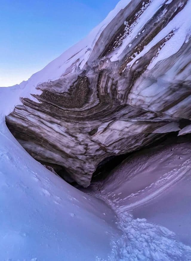 Primer plano de la entrada de una cueva de hielo jaspeada con capas de sedimento oscuro, rodeada de nieve profunda bajo un cielo despejado.