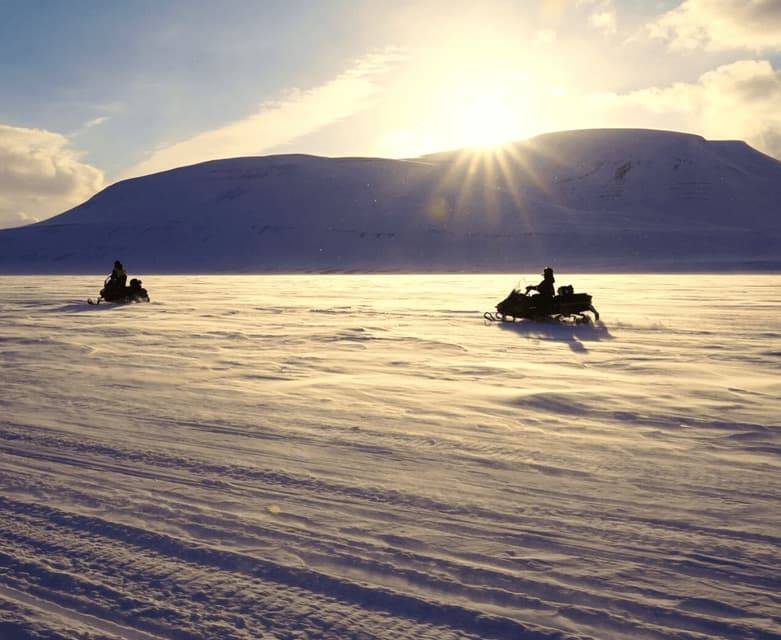 Un viaje en grupo de WeRoad en motos de nieve a través de una vasta, ventosa y nevada llanura mientras el sol se pone detrás de una montaña.