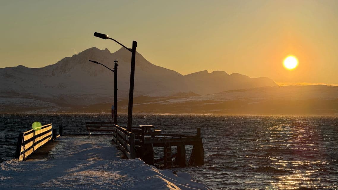 A snowy wooden pier extends into a body of water at sunset, with a backdrop of snow-covered mountains.