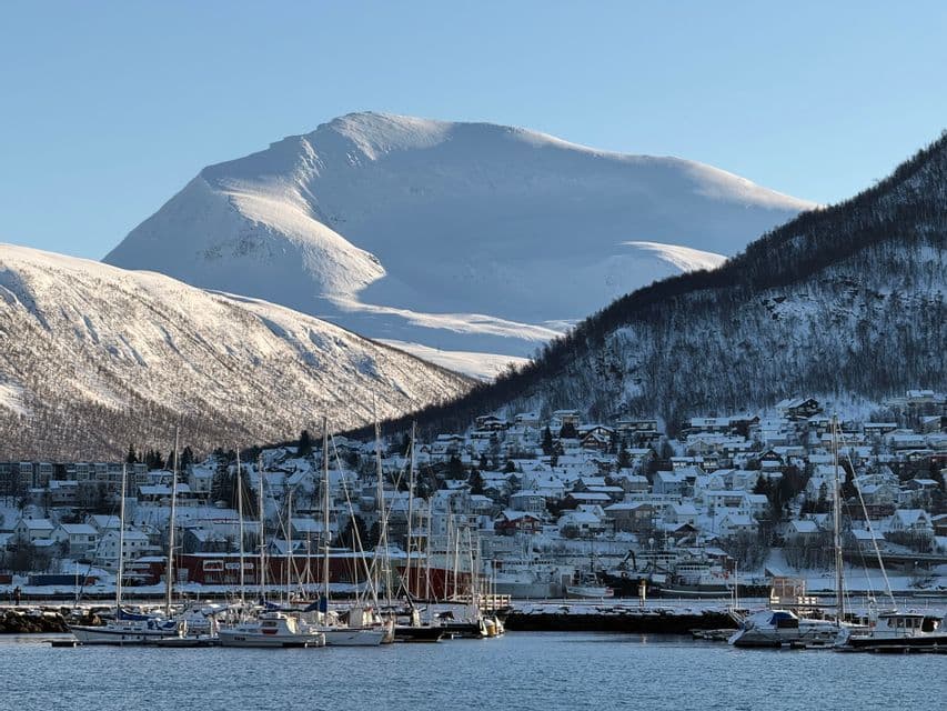 Sailboats are docked in a harbor in front of a snowy town nestled at the base of large, snow-covered mountains.